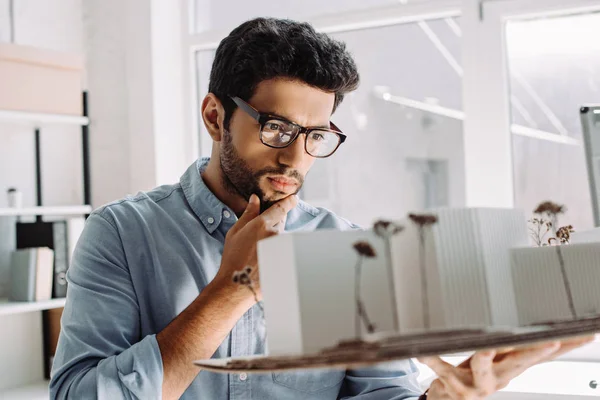 Arquitecto pensativo en gafas tocando barbilla y mirando el modelo de arquitectura en la oficina - foto de stock