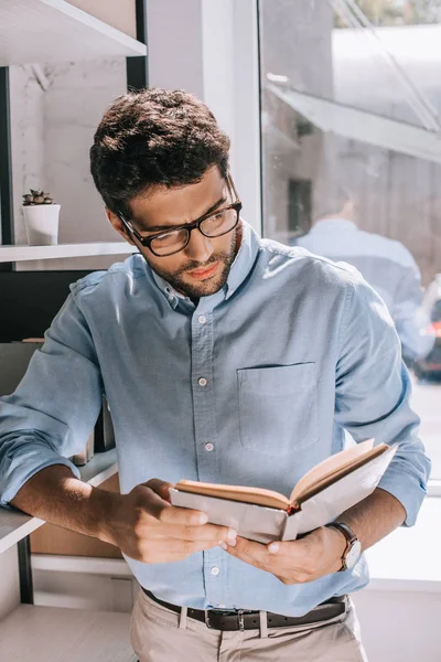 Handsome architect in glasses leaning on shelves and reading book in office — Stock Photo