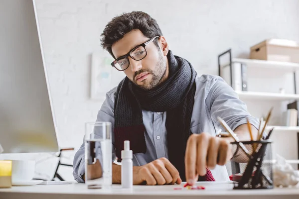 Sick businessman in scarf taking pill from pile in office — Stock Photo