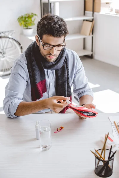 Sick manager in scarf holding first aid kit and electronic thermometer in office — Stock Photo