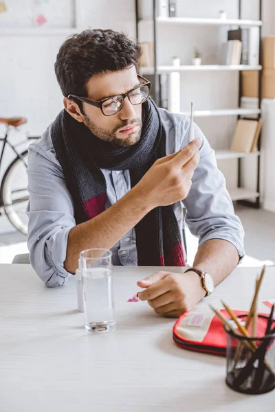 Sick manager in scarf looking at electronic thermometer in office — Stock Photo