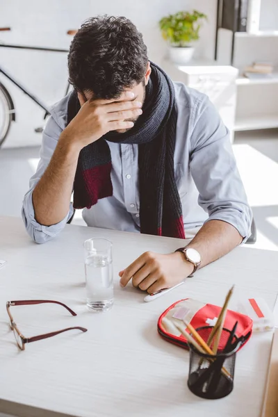 Hombre de negocios enfermo en bufanda tocando la frente y sentado en la mesa en la oficina - foto de stock