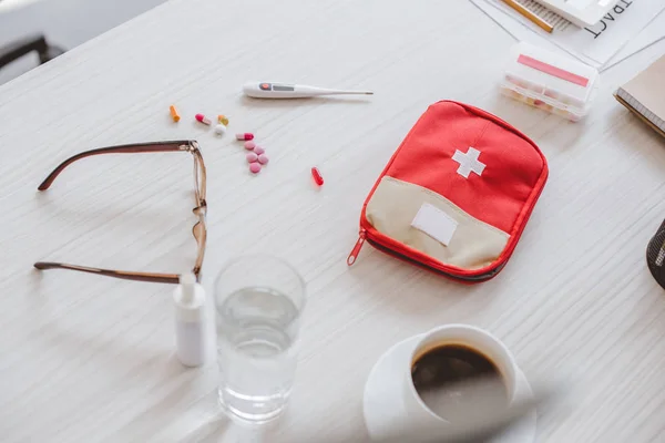 High angle view of first aid kit, glass of water, cup of coffee and pills on table in office — Stock Photo