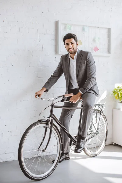 Homem de negócios sorridente sentado na bicicleta no escritório e olhando para a câmera — Fotografia de Stock