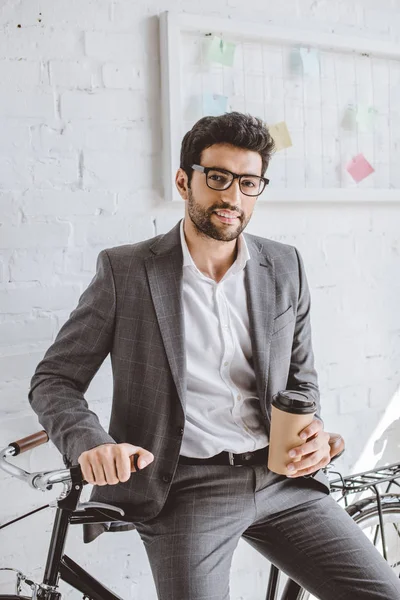 Smiling handsome businessman holding cup of coffee and leaning on bike in office — Stock Photo