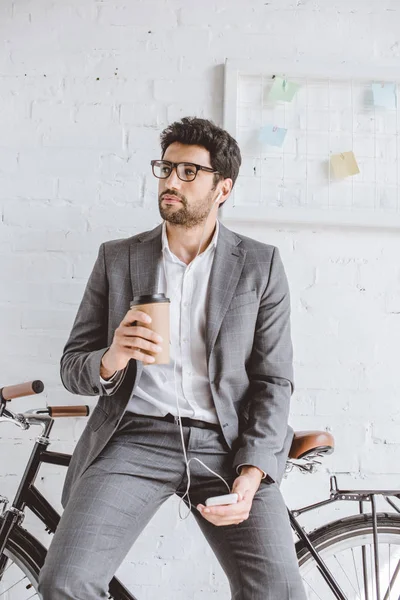 Uomo d'affari che ascolta musica con smartphone, tiene in mano una tazza di caffè e si appoggia alla bici in ufficio — Foto stock