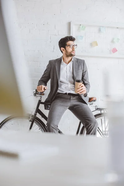 Bonito empresário sentado em bicicleta e segurando café para ir no escritório — Fotografia de Stock