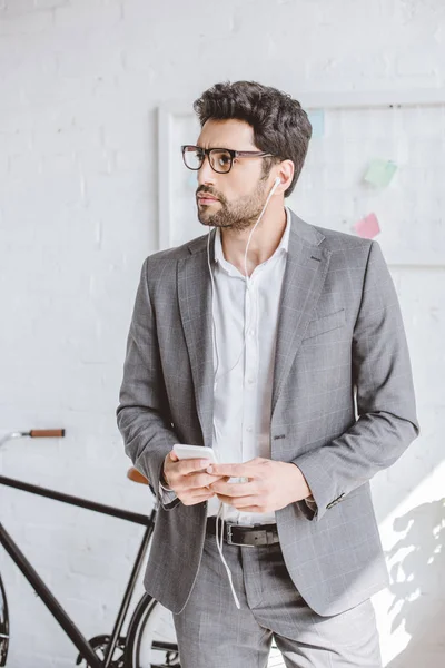 Hombre de negocios guapo escuchando música con teléfono inteligente y mirando hacia otro lado en la oficina - foto de stock