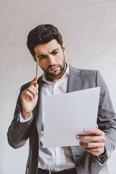 Homem de negócios sério lendo documentos e tocando a cabeça com lápis no escritório — Fotografia de Stock