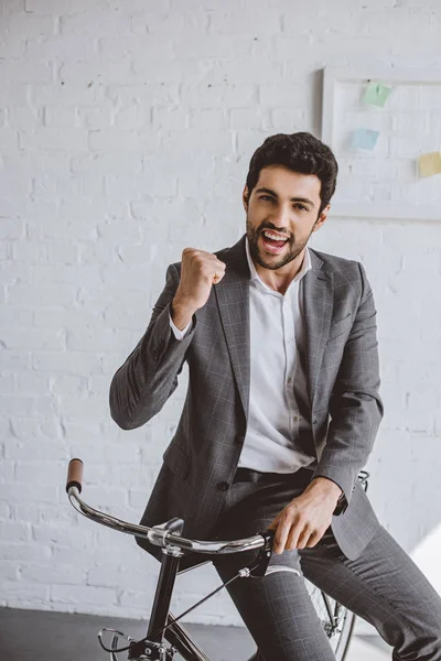 Homem de negócios bonito feliz sentado na bicicleta e mostrando sim gesto no escritório — Fotografia de Stock