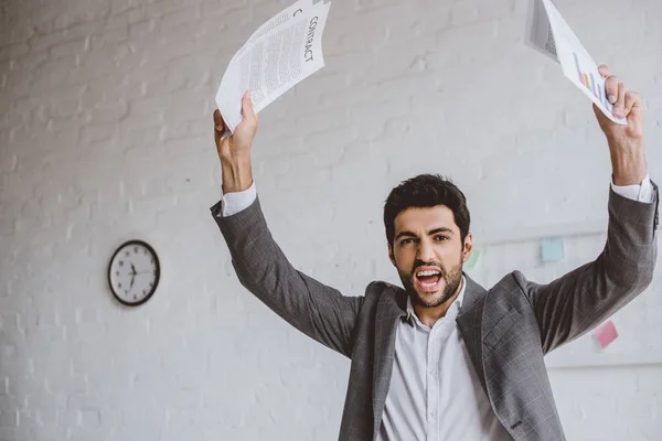 Excitado hombre de negocios guapo sosteniendo documentos en manos levantadas en la oficina - foto de stock