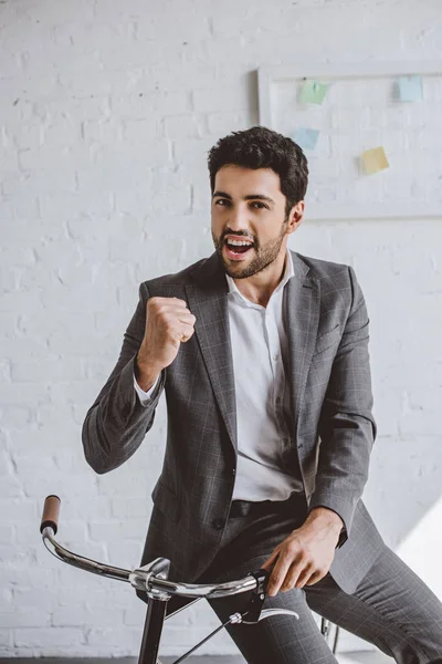Excited handsome businessman sitting on bike and showing yes gesture in office — Stock Photo
