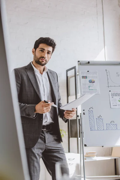 Guapo hombre de negocios sosteniendo documentos y rotulador cerca de rotafolio en la oficina — Stock Photo