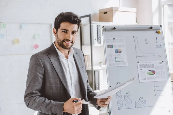 Smiling handsome businessman presenting project on flipchart and looking at camera in office — Stock Photo