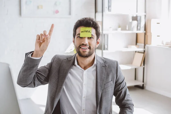 Alegre hombre de negocios apuntando hacia arriba y sentado con pegatina de papel en la frente con trabajo de palabra en la oficina - foto de stock