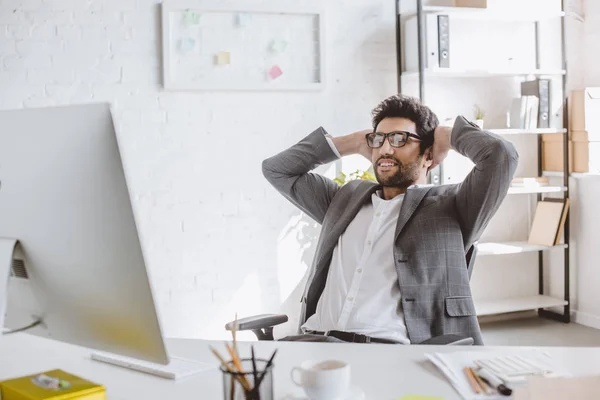 Glücklicher gutaussehender Geschäftsmann, der im Büro mit den Händen hinter dem Kopf auf den Computer schaut — Stockfoto