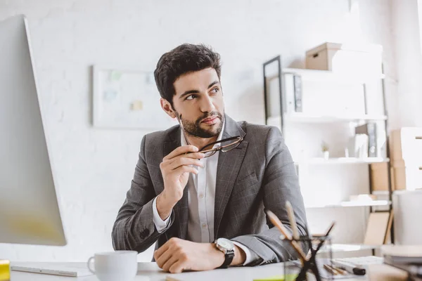 Handsome businessman holding glasses and looking away in office — Stock Photo