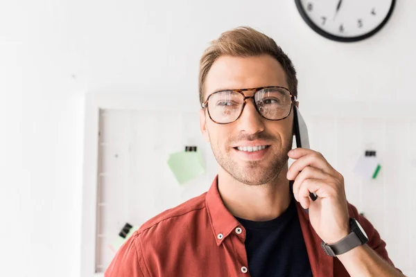 Улыбающийся бизнесмен в очках разговаривает на смартфоне в офисе — стоковое фото