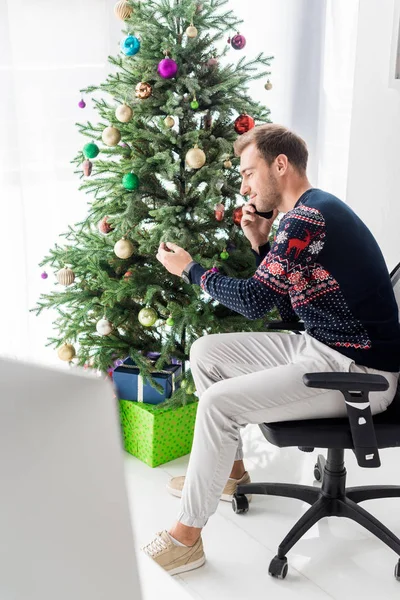 Mann im Weihnachtspulli sitzt auf Stuhl und telefoniert mit Smartphone neben Weihnachtsbaum — Stockfoto