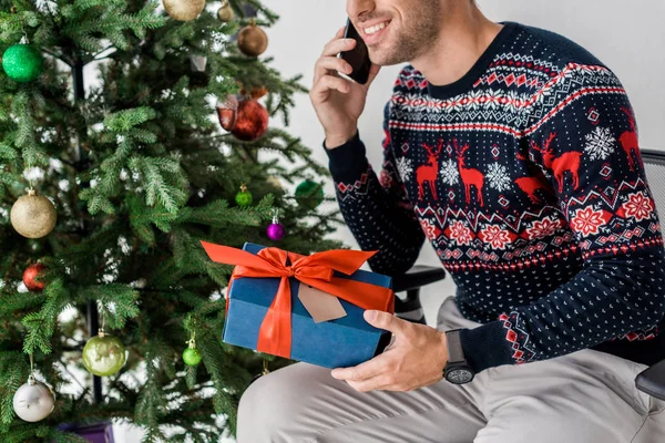 Hombre sonriente con regalo sentado cerca del árbol de Navidad y hablando en el teléfono inteligente - foto de stock
