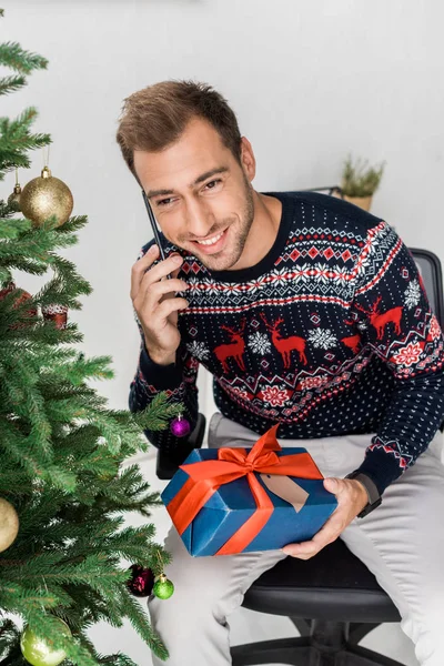 Hombre con regalo de Navidad sentado cerca del árbol de Navidad y hablando en el teléfono inteligente - foto de stock