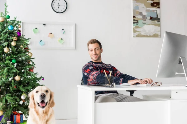 Lächelnder Mann im Weihnachtspulli sitzt am Tisch neben Golden Retriever Hund — Stockfoto