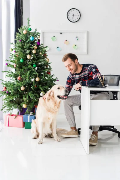 Man in christmas sweater sitting on chair and stroking golden retriever dog — Stock Photo