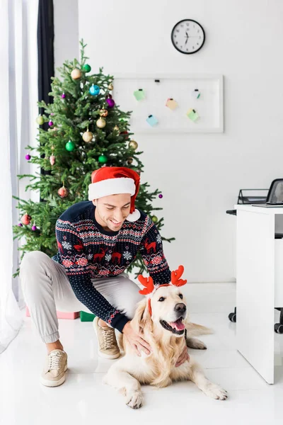 Улыбающийся мужчина в шляпе Санты гладит золотистую собаку-ретривер в оленьих рогах в канун Рождества — стоковое фото