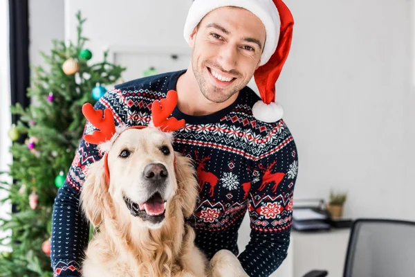 Man in christmas sweater and santa hat stroking dog in red deer horns — Stock Photo