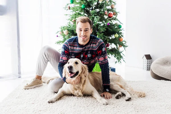 Happy man in sweater with golden retriever dog sitting near christmas tree — Stock Photo