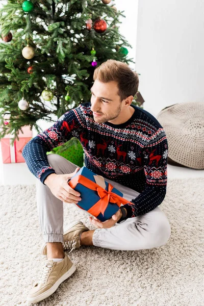 Mann im Weihnachtspullover mit Geschenkbox sitzt auf beigem Teppich — Stockfoto