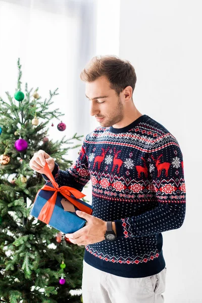 Mann im Weihnachtspullover öffnet Geschenkbox in der Nähe des Weihnachtsbaums — Stockfoto