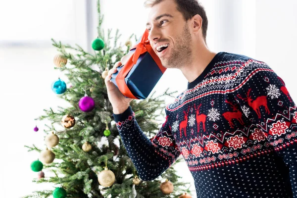 Man in christmas sweater with gift box near head against christmas tree — Stock Photo