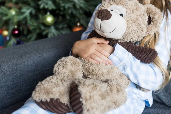 Обрезанный снимок ребенка, обнимающего плюшевого мишку и сидящего на диване на Рождество — стоковое фото