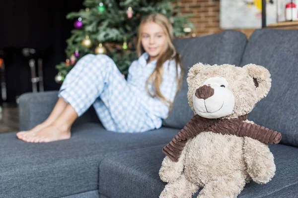 Крупный план плюшевого мишки и ребенка в пижаме, сидящего на диване позади на Рождество — стоковое фото