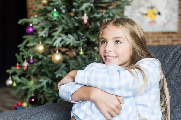 Criança feliz bonito em pijama sentado no sofá e olhando para longe no tempo de Natal — Fotografia de Stock