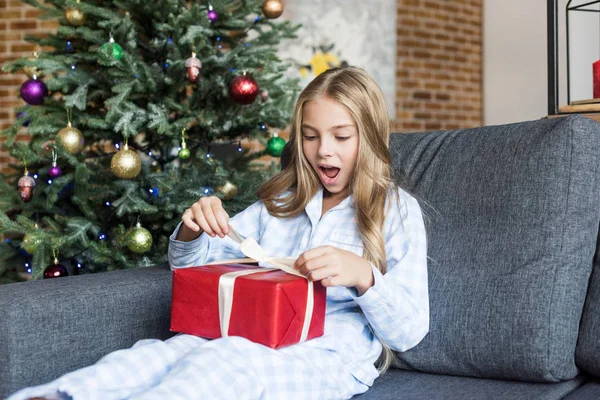 Niño sorprendido en pijama abriendo regalo de Navidad mientras está sentado en el sofá - foto de stock