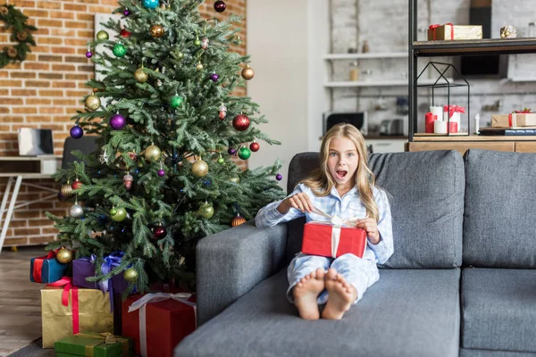 Criança surpresa em pijama abrindo presente de Natal e olhando para a câmera — Fotografia de Stock