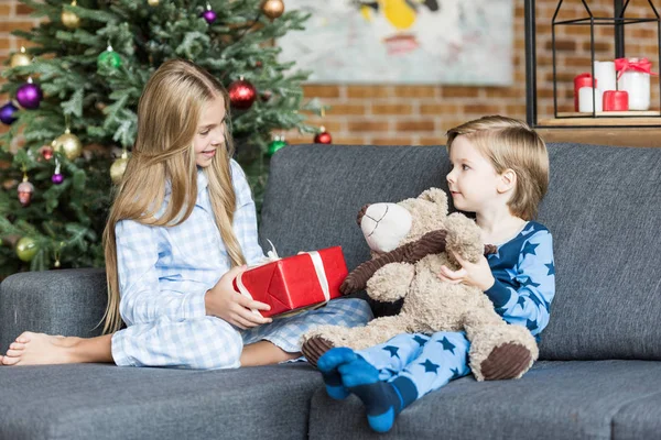 Cute happy kids in pajamas holding teddy bear and christmas present while sitting in sofa and smiling each other — Stock Photo