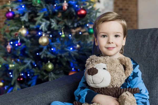 Adorável criança feliz abraçando ursinho de pelúcia e sorrindo para a câmera enquanto sentado no sofá no tempo de Natal — Fotografia de Stock