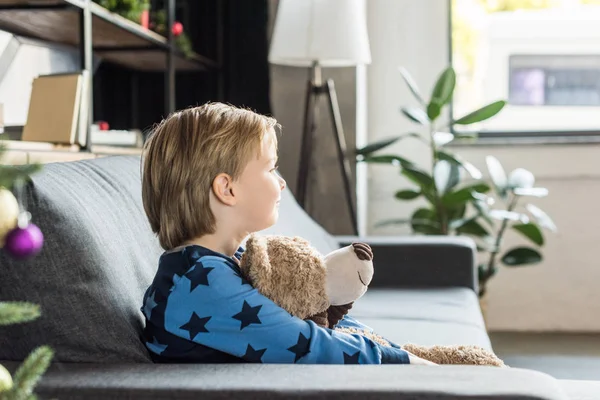 Vista lateral da criança feliz adorável segurando ursinho de pelúcia e olhando para longe enquanto sentado no sofá — Fotografia de Stock