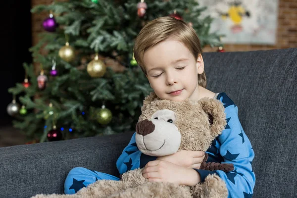 Lindo niño pequeño en pijama abrazando oso de peluche mientras duerme y se sienta en el sofá - foto de stock