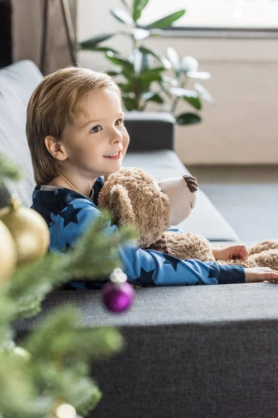 Vista lateral de lindo niño sonriente sosteniendo oso de peluche y sofá sentado - foto de stock