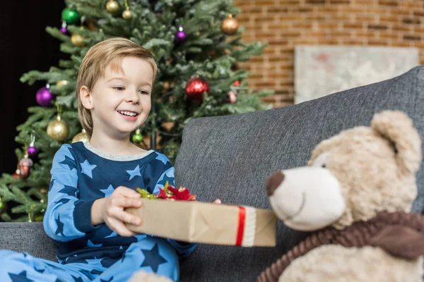 Mignon enfant heureux en pyjama tenant cadeau de Noël et regardant ours en peluche — Photo de stock