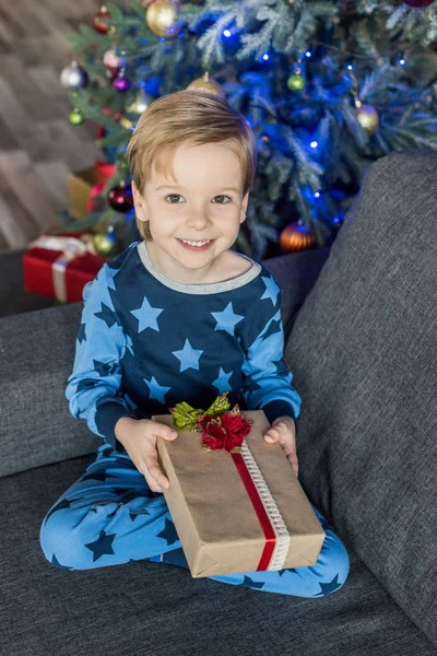 Entzückendes glückliches Kind im Pyjama mit Weihnachtsgeschenk und lächelnd in die Kamera — Stockfoto
