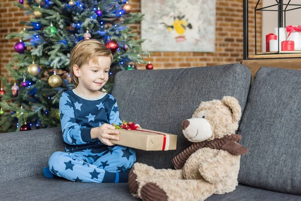 Очаровательный ребенок в пижаме с рождественским подарком и глядя на плюшевого мишку — стоковое фото