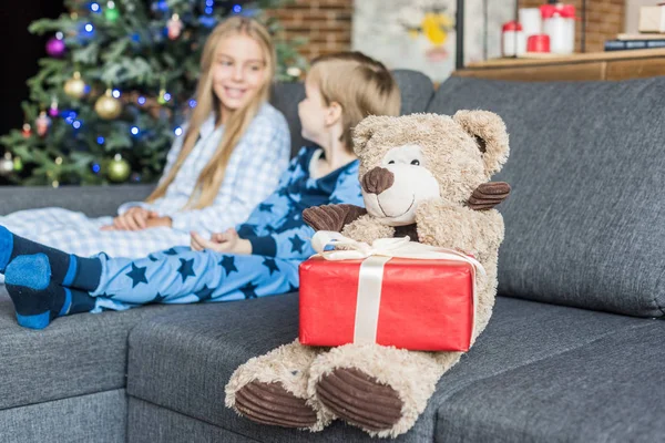 Vista de cerca del oso de peluche con regalo de Navidad y los niños en pijama sentados en el sofá detrás - foto de stock