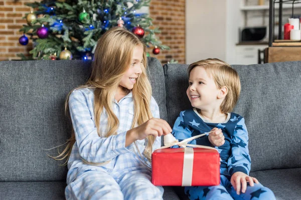 Niedliche glückliche Kinder im Pyjama halten Weihnachtsgeschenk und lächeln einander an — Stockfoto