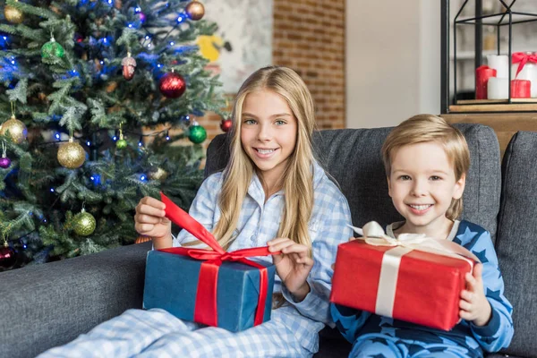 Lindos niños felices en pijamas sosteniendo regalos y sonriendo a la cámara en Navidad - foto de stock