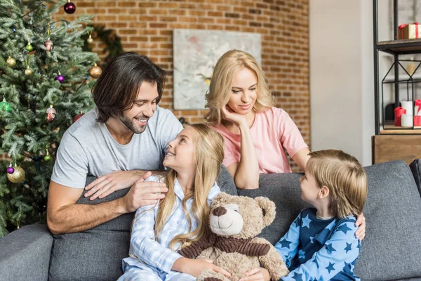 Happy parents looking at adorable smiling children in pajamas sitting in sofa at christmas time — Stock Photo